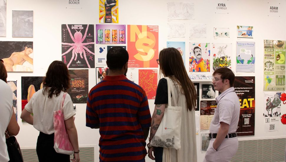 A group of people viewing student artwork displayed on a gallery wall, featuring colorful posters, paintings, and graphic designs at the AFA Graduation Exhibition at Delaware County Community College.