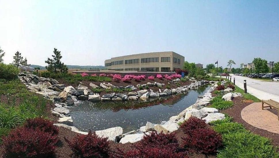 The campus at Penn State Great Valley in Malvern from afar on a sunny day. There is a small water installment/pond surrounded by rocks and lush grass and greenery and budding flowers.