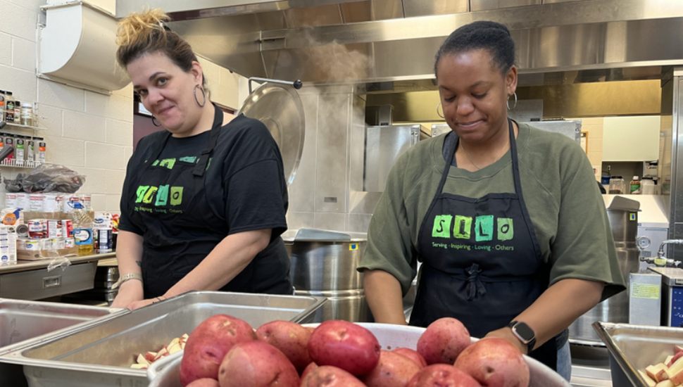 Two women volunteers at SILO prepare food for clients in a kitchen, with a bowl of tomatoes nearby.