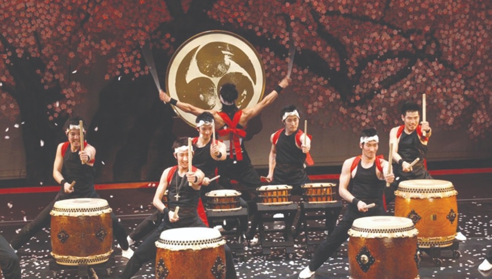 A dynamic performance by Tamagawa University Taiko drummers and dancers, dressed in black and red traditional attire, energetically playing large taiko drums on stage against a backdrop of cherry blossoms and a large Japanese emblem.