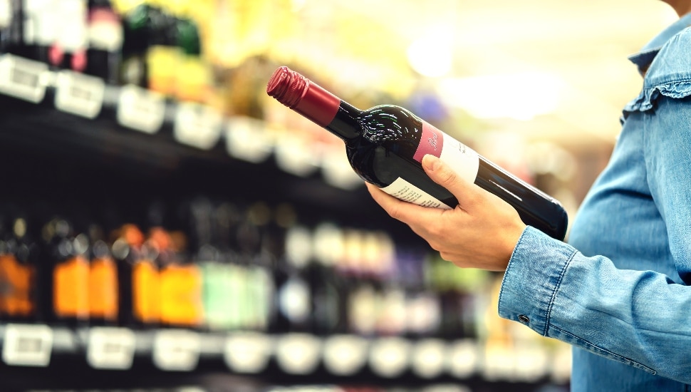 A woman holds a bottle of wine in a liquor store with shelves full of liquor bottle in front of him.