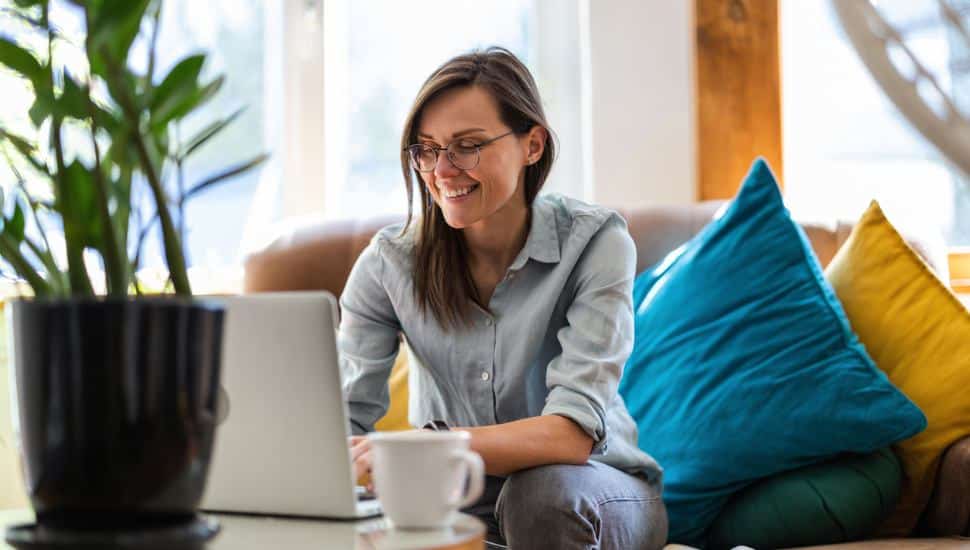 A woman happily works at her computer sitting on a sofa drinking coffee.