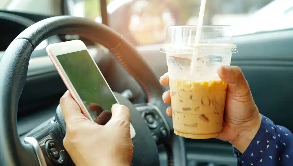 A driver behind the wheel of a car holding an iced coffee and looking at their mobile phone.