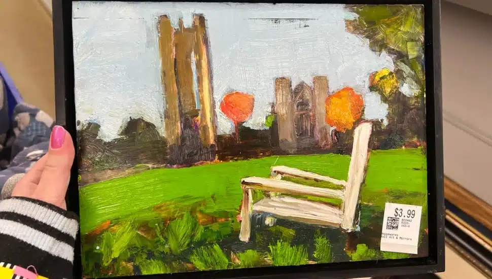 A woman holds a painting of a white bench against a green lawn and stone tower she picked up in a thrift store.
