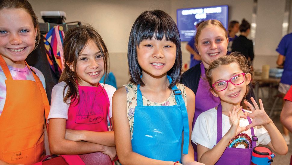 Group of smiling young girls wearing colorful aprons at a summer kids camp, posing together indoors during a cooking or creative activity.