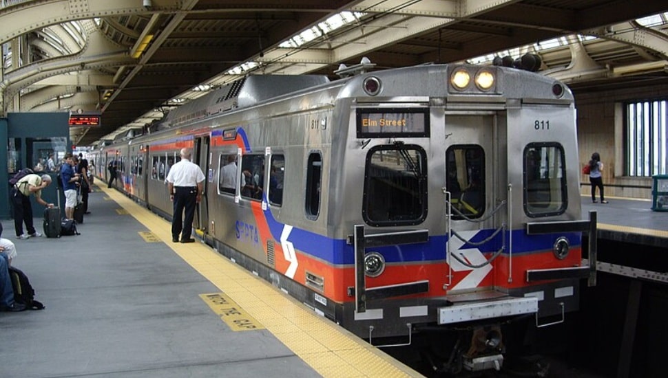 A regional rail train is stopped at a station as passengers stand on the platform and a conductor stands by an open train door.