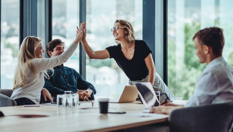 Four employees at a table, one with a laptop. Two employees are congratulating each other with a high-five.
