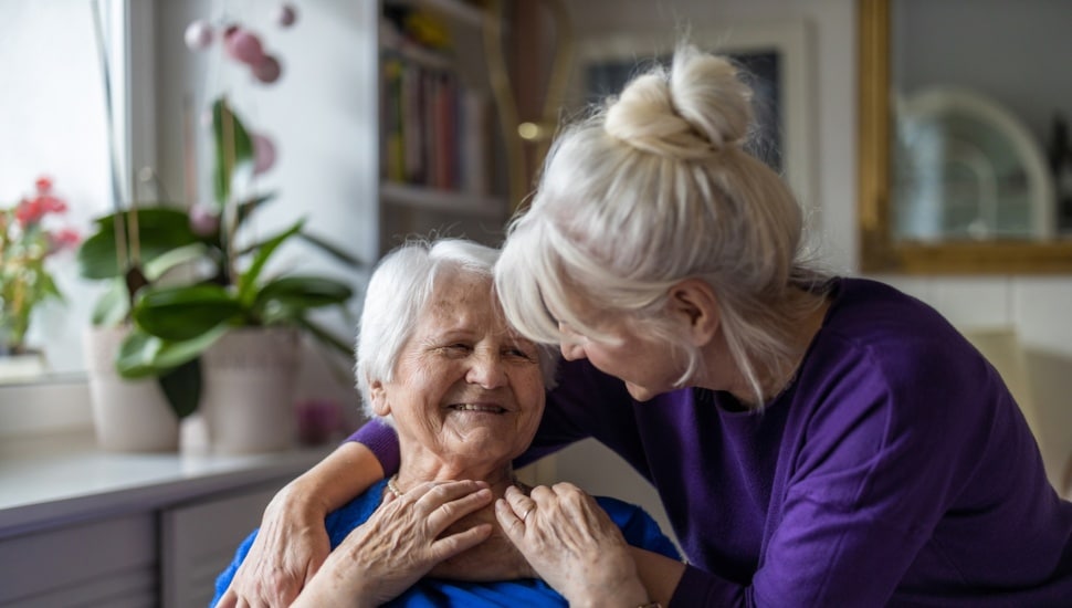 A woman comforts an elderly woman sitting in a chair.