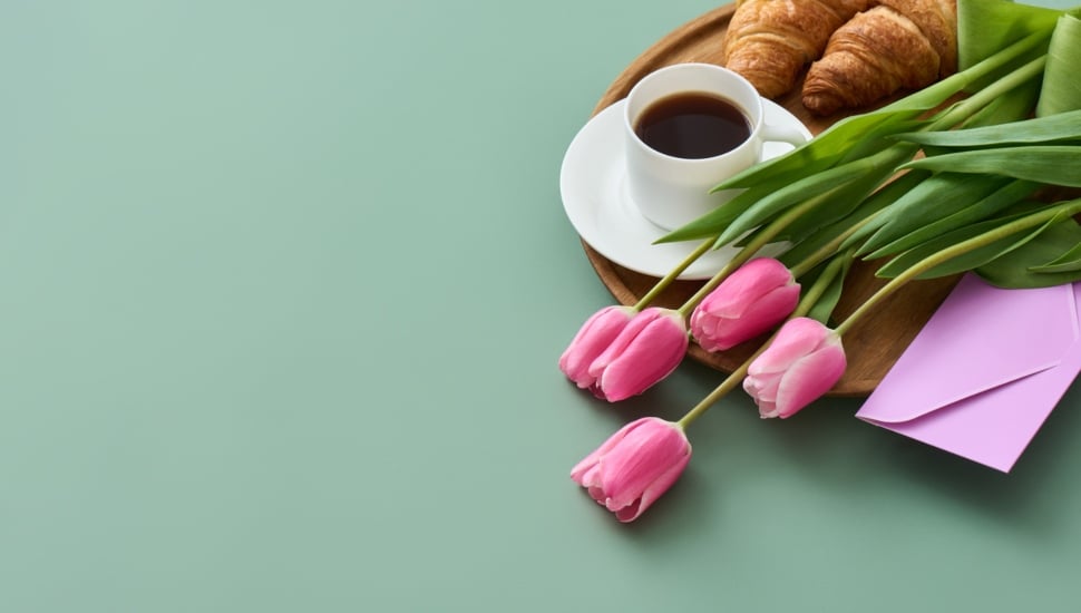 Cut flowers and a card lie on a plate that has coffee and a croissant for Mother's Day.