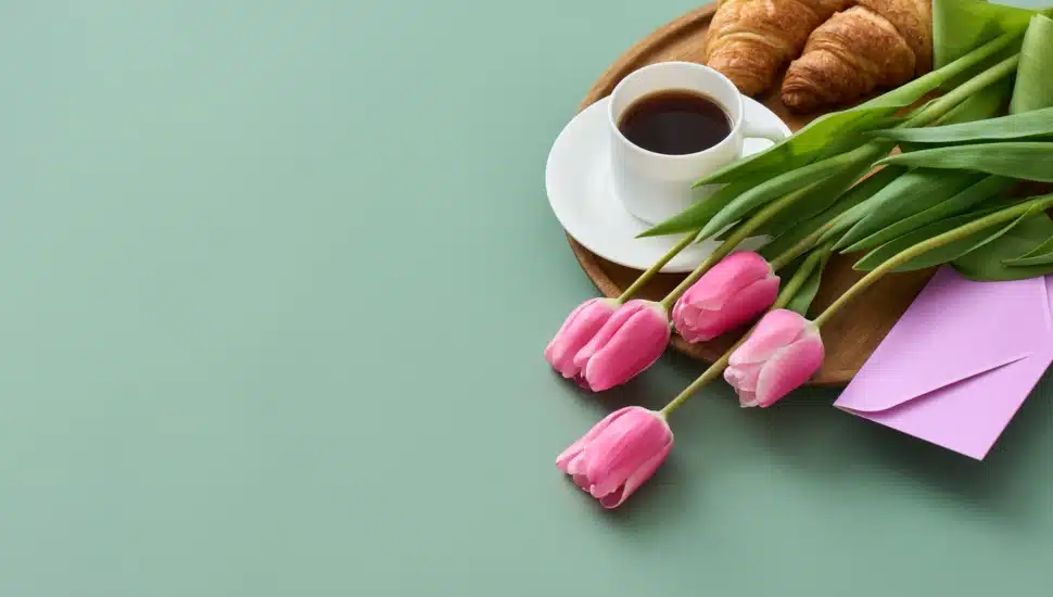 Cut flowers and a card lie on a plate that has coffee and a croissant for Mother's Day.