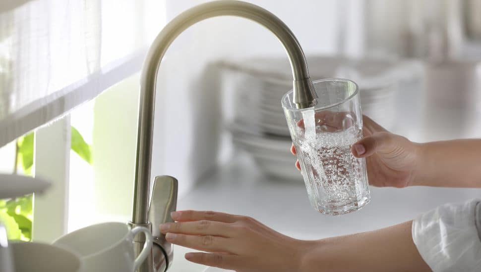 Person filling a clear glass with water from a modern kitchen faucet in a brightly lit kitchen.