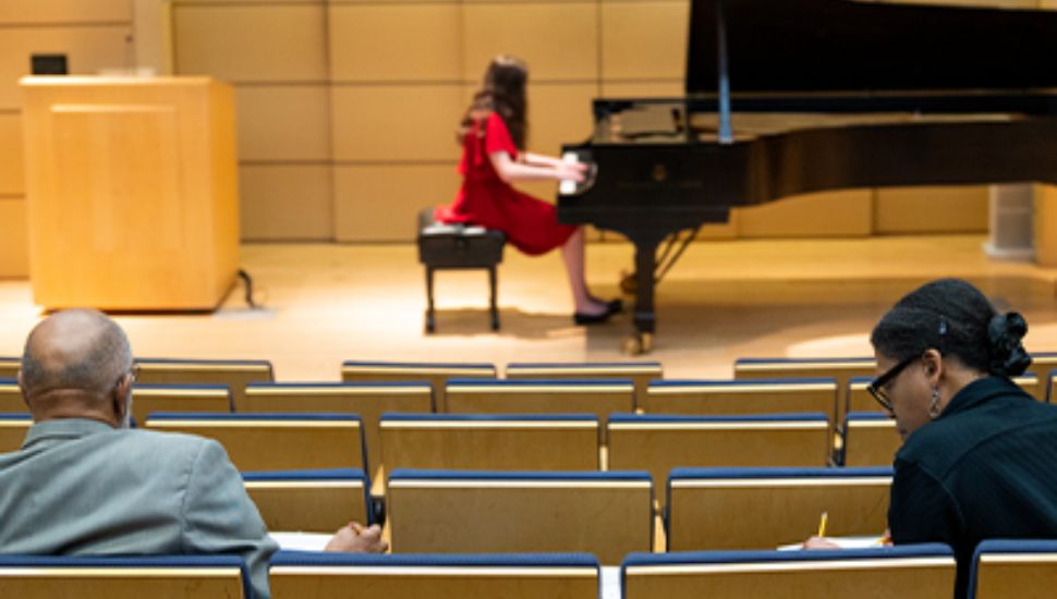 A girl with long brown hair and a red dress playing the piano at a distance on stage. Judges sit in seats taking notes.
