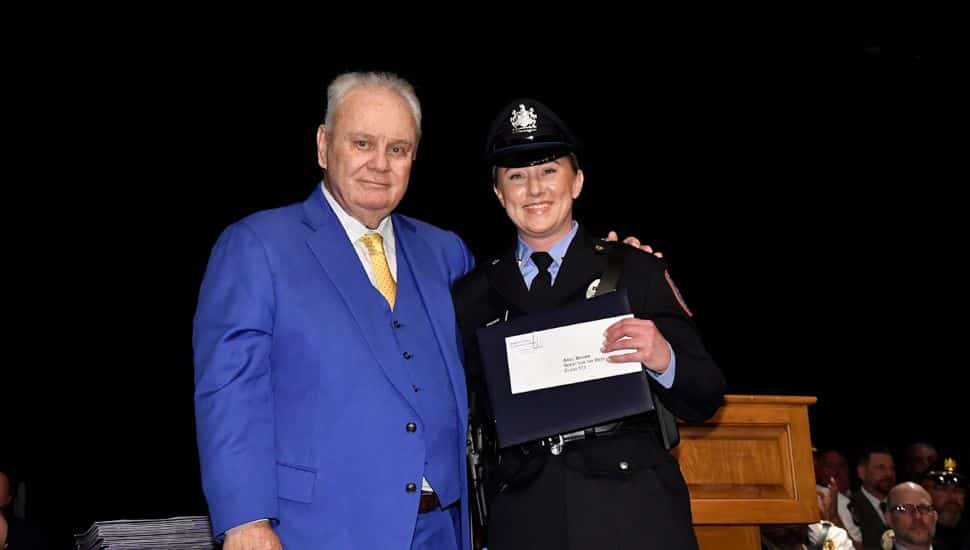 MPA Director William Davis with Cadet Ariel Brown of Philadelphia. Police academy graduate in uniform smiles while holding a certificate, standing next to a man in a blue suit during a graduation ceremony.              
