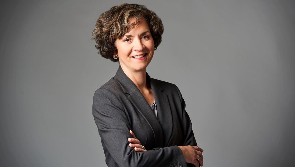 Dr. Marta Yera Cronin, President of Delaware County Community College, poses confidently in a professional dark gray suit against a plain gray background, smiling with arms crossed.