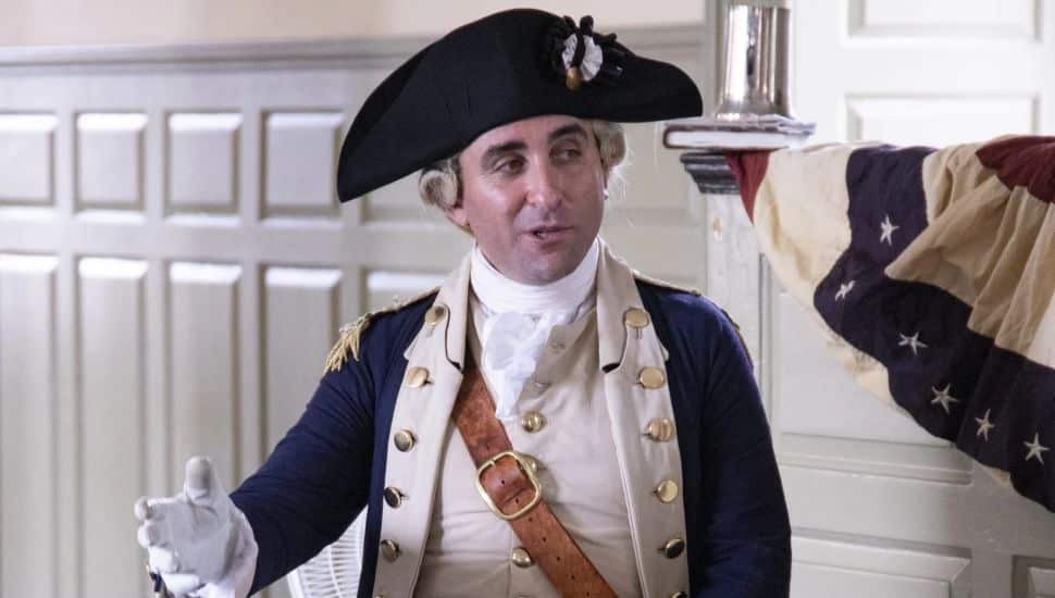 A historical interpreter dressed as Marquis de Lafayette wears an 18th-century Continental Army uniform with a navy blue coat, white waistcoat, and a black tricorn hat. He gestures with one hand while seated indoors beside a draped colonial American flag.