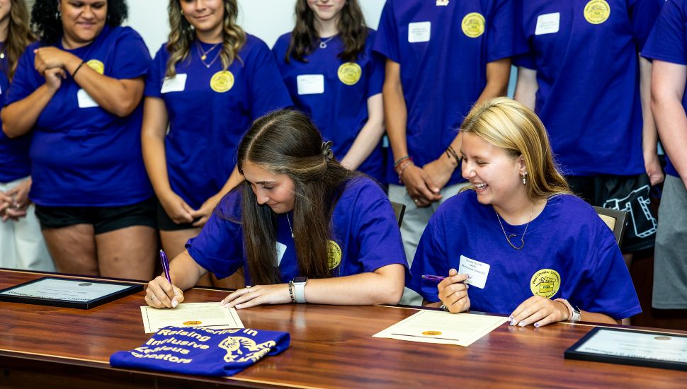 (L to R) During the 2024 inaugural signing ceremony, WCU PRIZE students Hannah Giampietro and Cecelia Perrotti (both from Kennett High School) pledged “unwavering zeal” to the students they will teach one day: Two high school seniors, wearing purple PRIZE Program t-shirts with gold emblems, sit at a wooden table smiling as they sign official commitment declarations to become future educators. Around them, a group of supportive peers—also in matching PRIZE shirts—stand watching the ceremony. The scene takes place during West Chester University’s inaugural PRIZE Signing Day, part of an initiative to address the national teacher shortage by nurturing future teachers from local school districts.