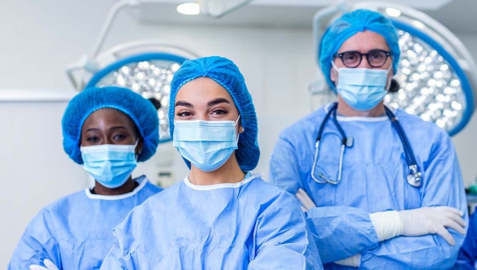 Three medical professionals wearing blue surgical scrubs, masks, gloves, and hair coverings stand confidently in an operating room. The person in the center is smiling behind the mask, with surgical lights visible in the background.