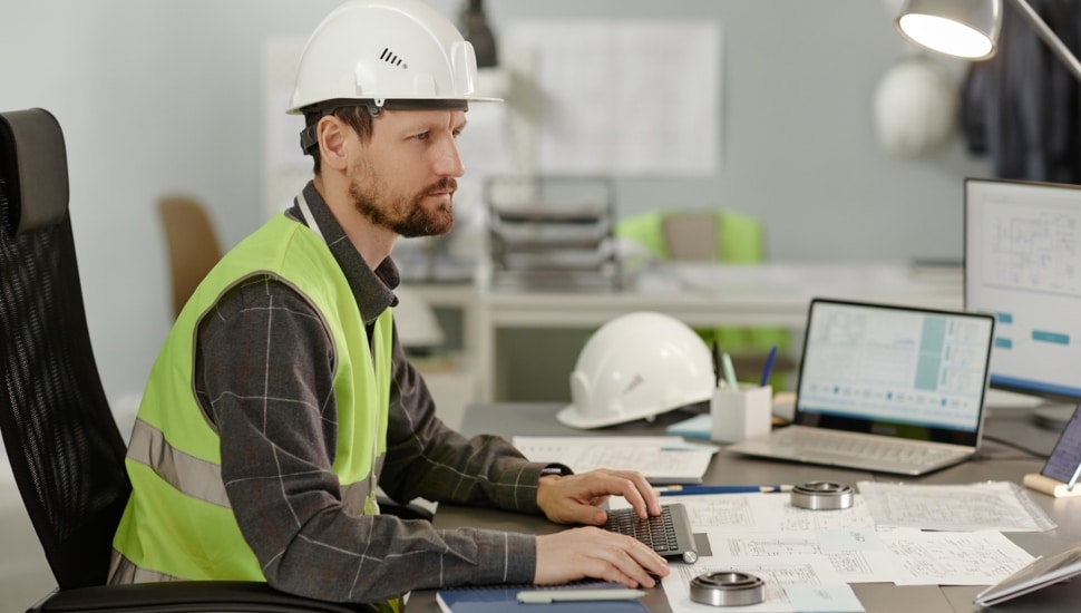 An engineer wearing a construction hat sitting at his desk working at a keyboard.