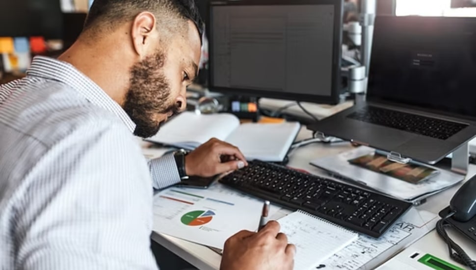 A man in a lab coat writing something down next to a computer terminal.