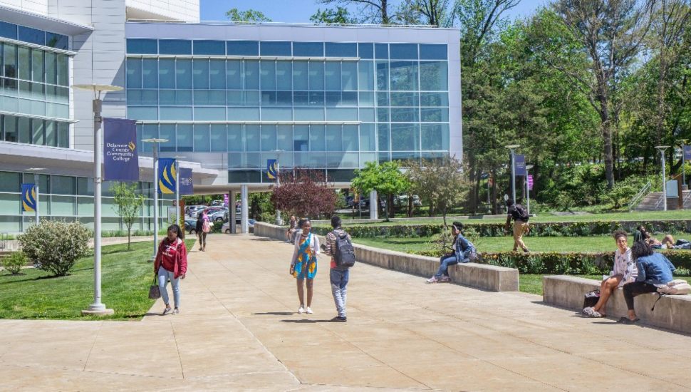 Students walk and relax outside on a sunny day at Delaware County Community College’s main campus. The scene shows a modern glass academic building, landscaped pathways, and students enjoying the green campus environment.