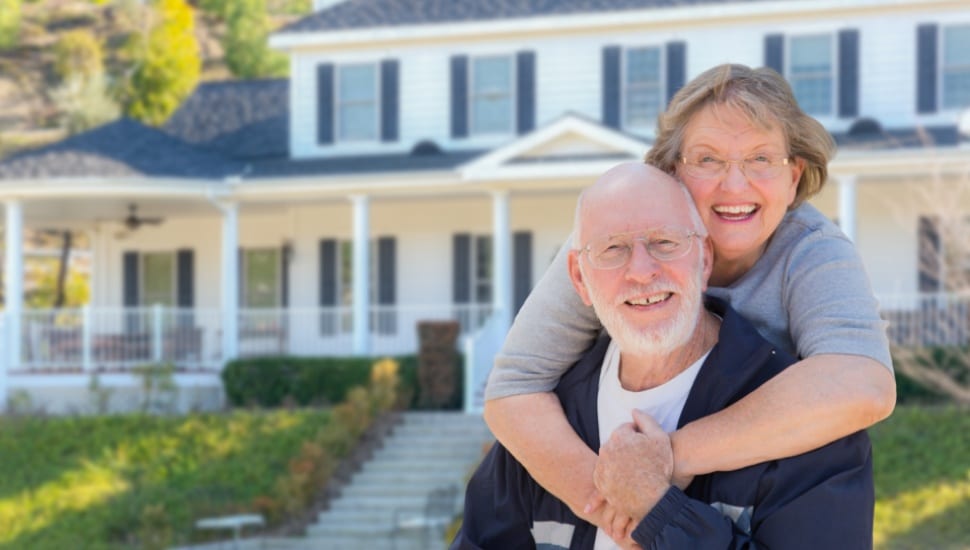 A senior couple embrace in front of their home.