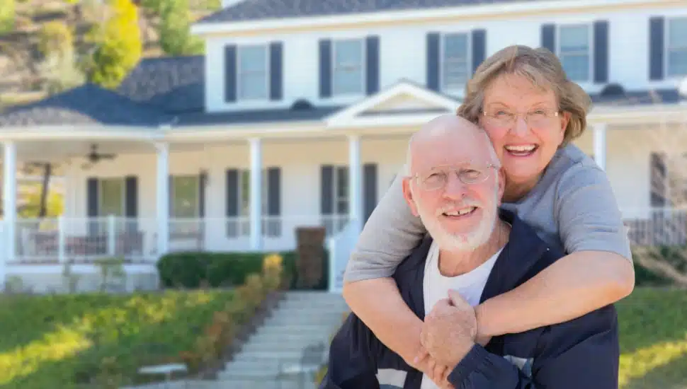 A senior couple embrace in front of their home.