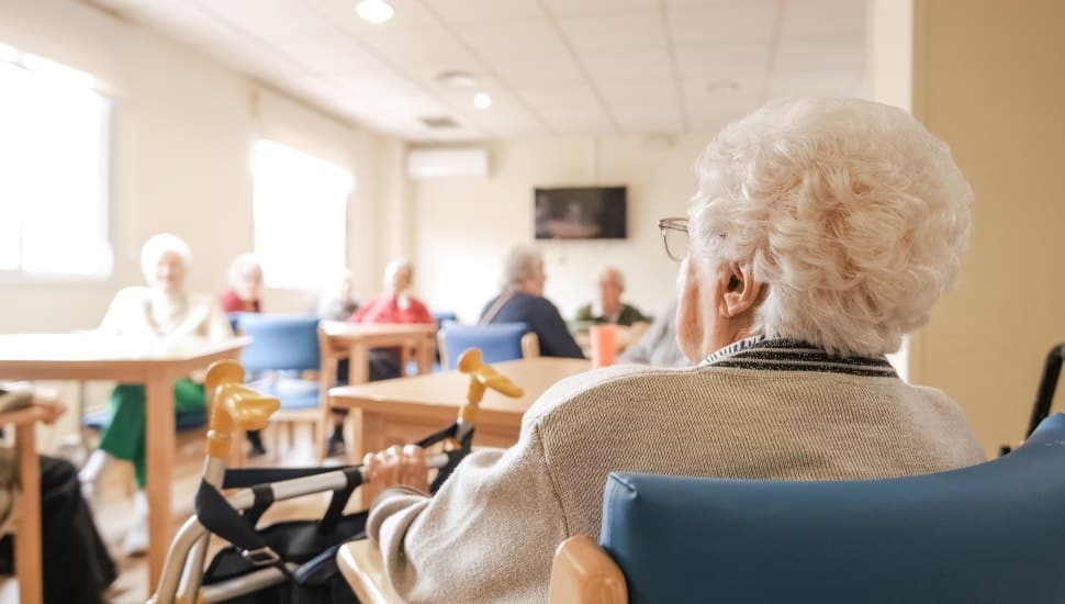 An elderly woman sits in a room at a nursing home.