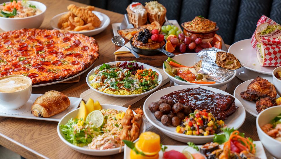 A vibrant spread of global dishes at Southern Market in Lancaster, featuring pepperoni pizza, BBQ ribs, colorful rice bowls, grilled meats, fresh fruit, wraps, pastries, and coffee — all beautifully arranged on a wooden table.
