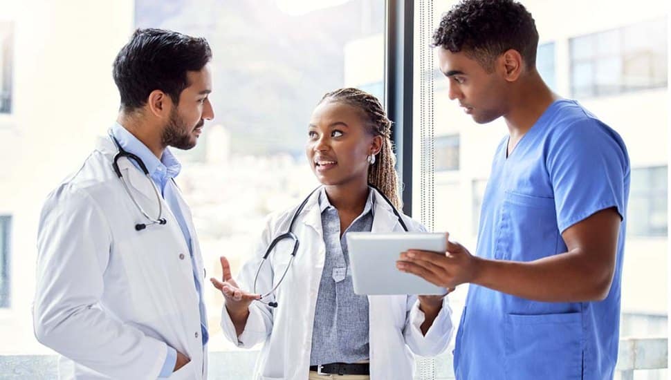 Three medical professionals, two in white lab coats and one in blue scrubs, are standing near a window in a hospital setting. They are engaged in a focused and collaborative conversation. One person holds a tablet, suggesting they are reviewing medical information or patient data. The atmosphere appears professional and cooperative.