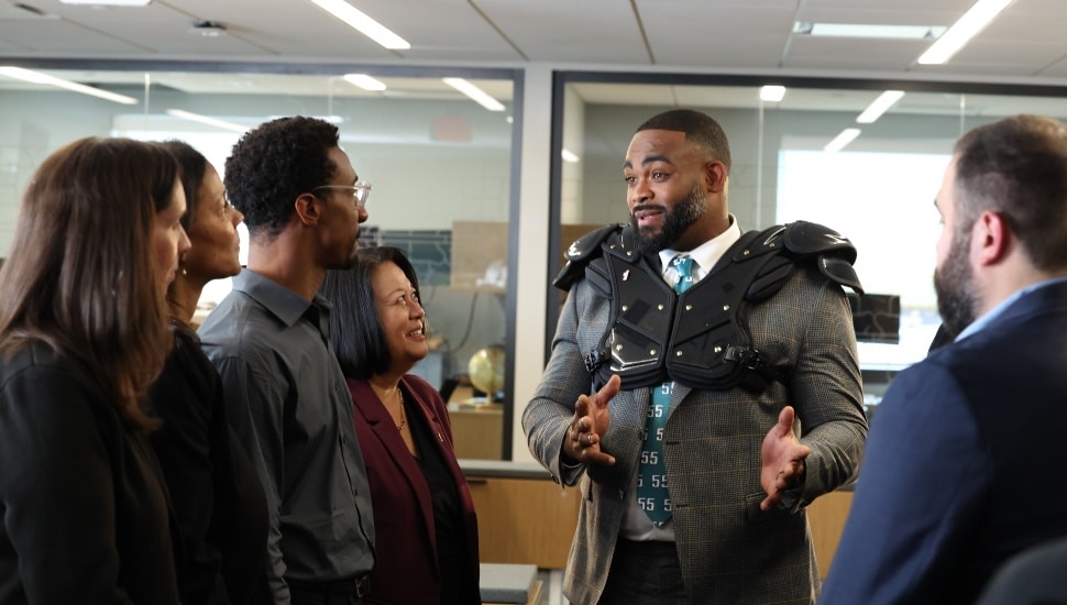 Football player wearing suit and shoulder pads talks with bank employees.