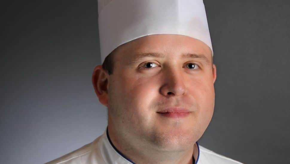 Portrait of Chef Jonathan Fike wearing a traditional white chef's uniform and tall pleated chef hat. His coat is embroidered with his name and professional titles: “Jonathan Fike, CEC, CCA.” He stands against a neutral gray background, looking confidently into the camera.