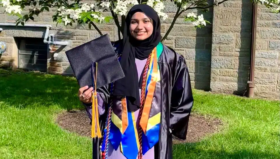 Needah-e-Fatema Hafeezali Gangji, wearing a graduation cap and gown with honor cords and stoles, smiling outdoors on the Delaware County Community College campus.