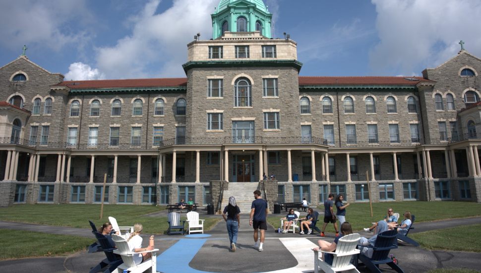 Front view of Immaculata University’s historic main building with its stone facade and green-domed tower, as students walk, sit in Adirondack chairs, and socialize on the lawn under a partly cloudy sky.