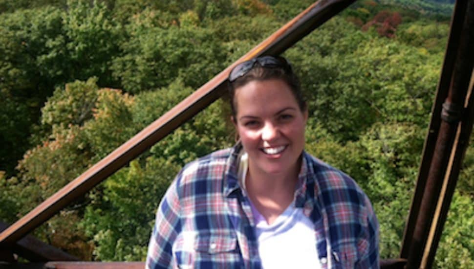 Smiling woman in plaid shirt standing on a metal structure with green trees in the background.