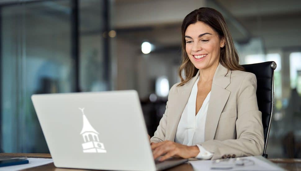 Professional woman smiling while working on a laptop with Wilmington University’s tower logo in a modern office setting.
