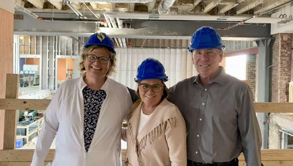 Three people wearing blue hard hats stand inside a construction site. From left to right: Dr. Marian Kozachyn, Vice President at DCCC, Sharon Kelleher, and Andrew Kelleher. They are smiling while visiting the future Southeast Center in Drexel Hill, PA.