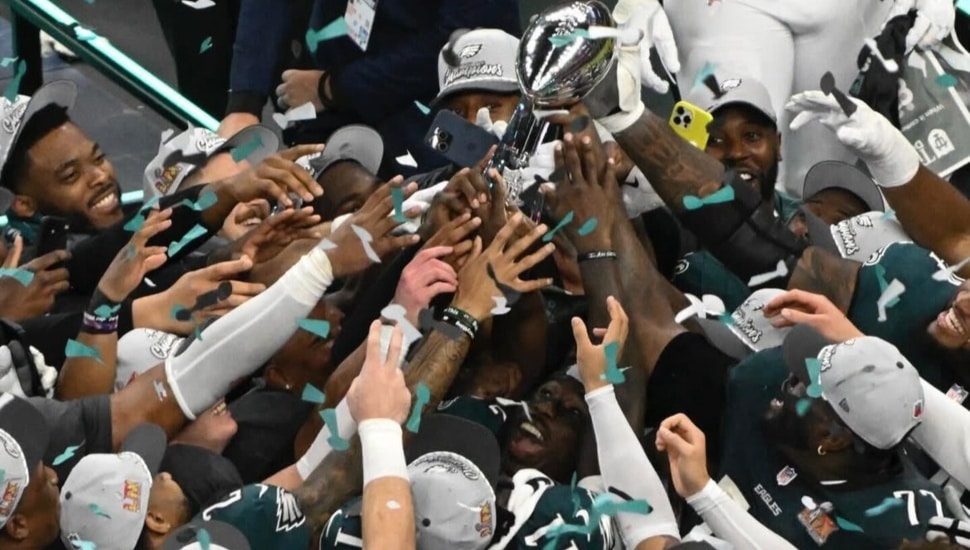 A crowd gathers around the Lombardi Trophy, each person trying to touch it after an Eagles Super Bowl win.