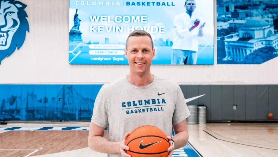Male college basketball coach poses for picture in gym with basketball in hand.