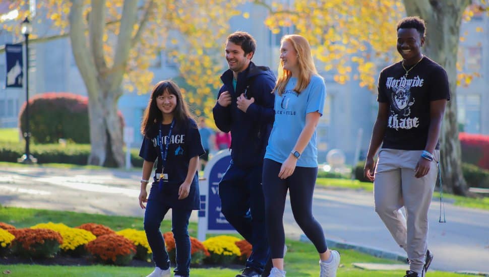 Four Immaculata University students walking together on campus during fall, smiling and talking near colorful autumn trees and flowers.