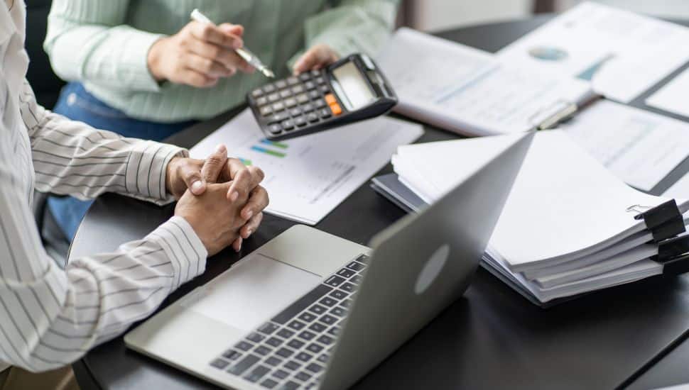 Two people are sitting at a desk using a calculator and filling out tax forms.