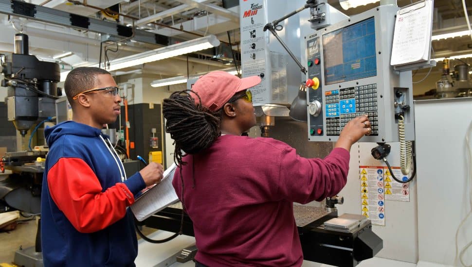 Two Delaware County Community College students operate an industrial training machine in a lab, learning mechanical and electrical system controls as part of a workforce development program.