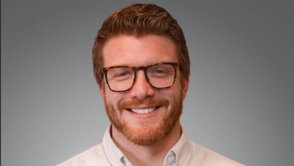 A man with short, wavy red hair, a beard, and glasses smiling at the camera against a gray background.