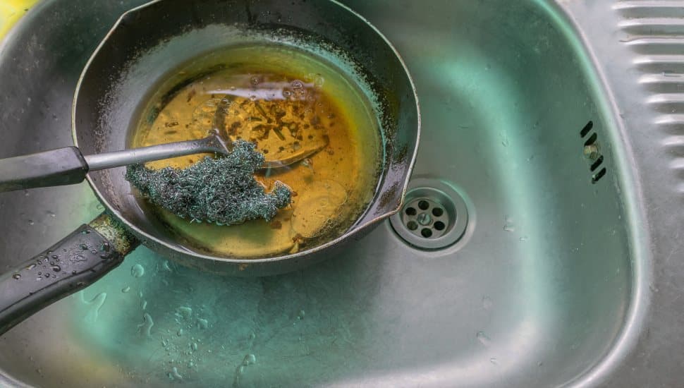 A frying pan filled with leftover cooking oil sits in a kitchen sink, with a metal scrubber resting inside the pan. Water droplets are visible around the stainless steel sink, highlighting the mess left after cooking.