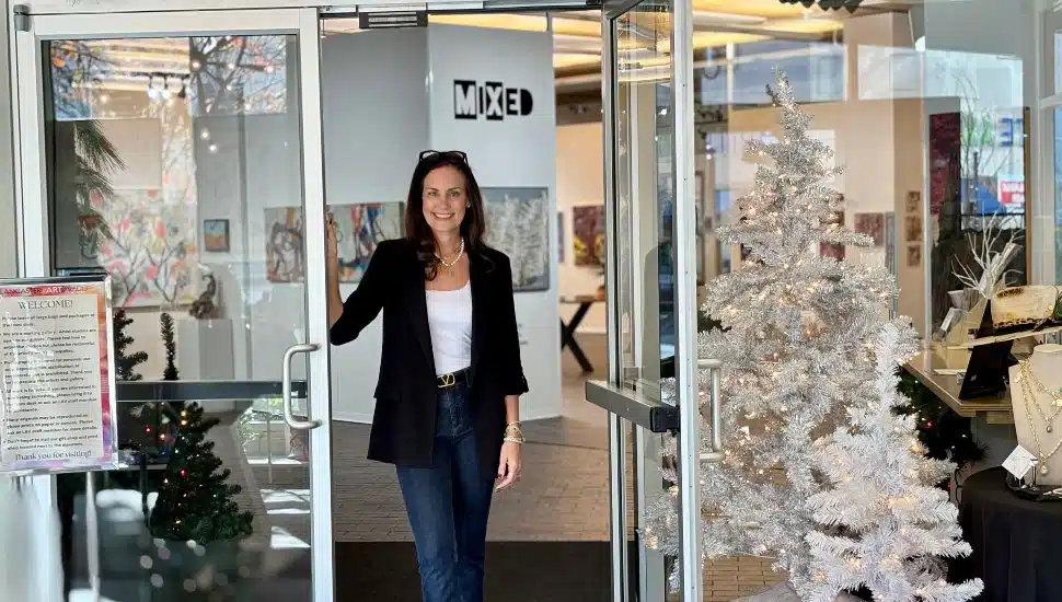 A woman stands smiling in the open doorway of the Lancaster Art Vault gallery, surrounded by holiday decorations and illuminated displays of artwork inside.