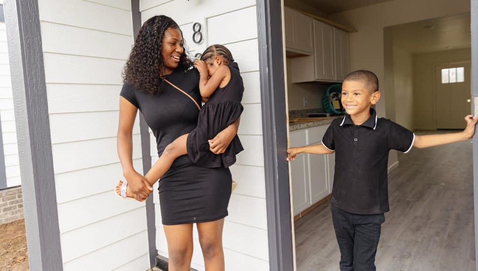 A young mother holds her daughter next to her son in front of their new home.
