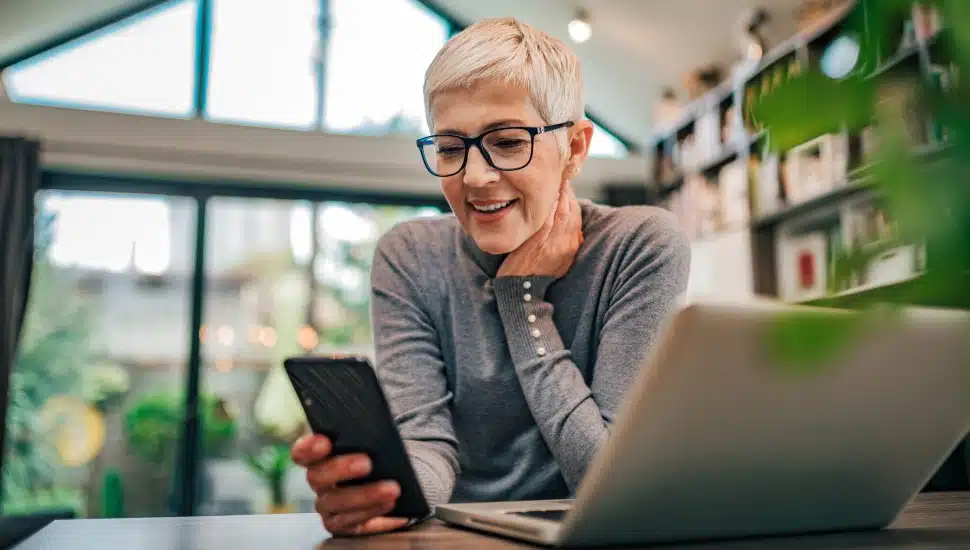 A senior woman sits at home looking at her laptop and smart phone.