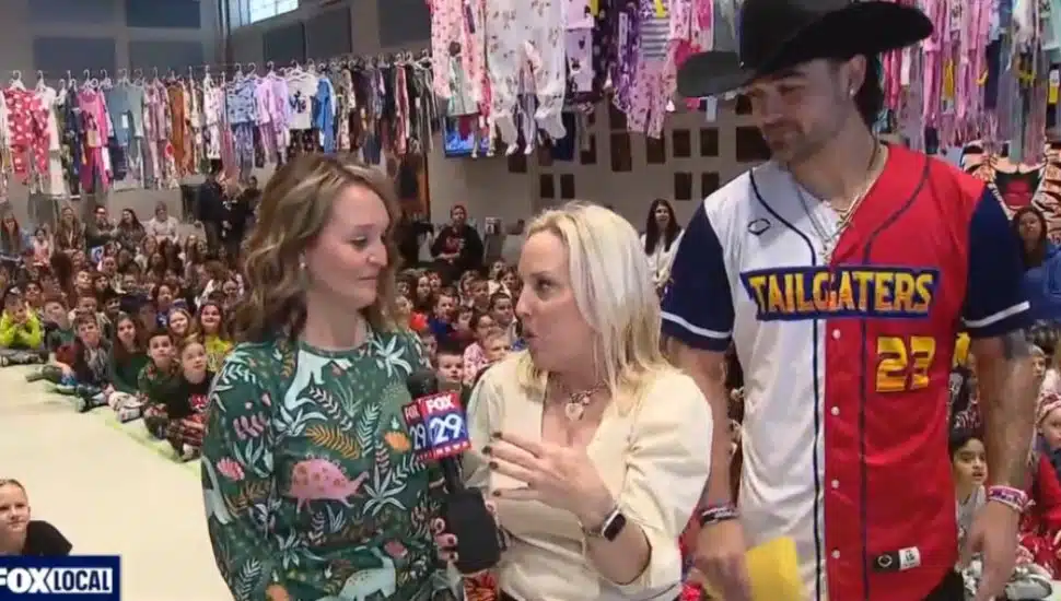Fox 29 reporter Jenn Frederick talks with Jaime Smyth , principal at Loomis Elementary School and Joe Sperone, a pitcher with the Texas Tailgaters. Collected pajamas hang from rafters in the background while students sit on the floor.