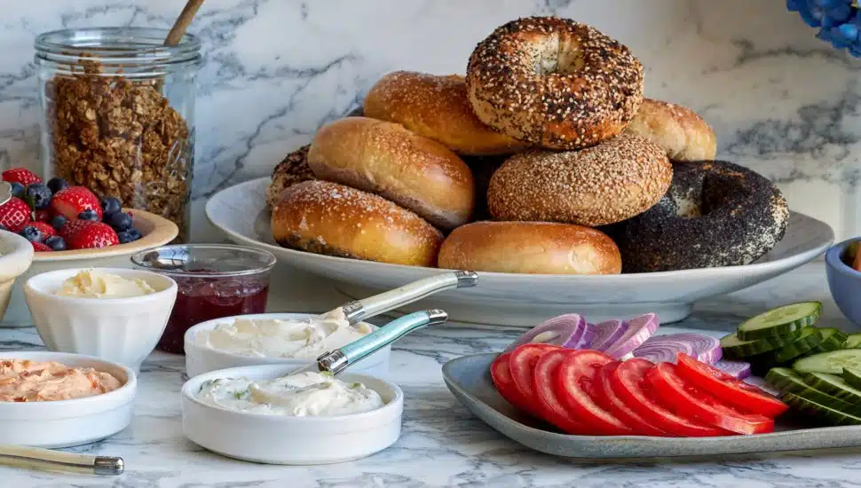A platter of fresh bagels with different spreads, tomato and cucumber slices, and fresh strawberries.