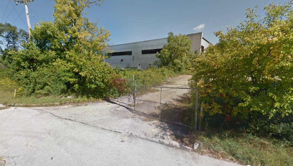 Former Bishop Tube site with overgrowth of plants and fence blocking driveway.