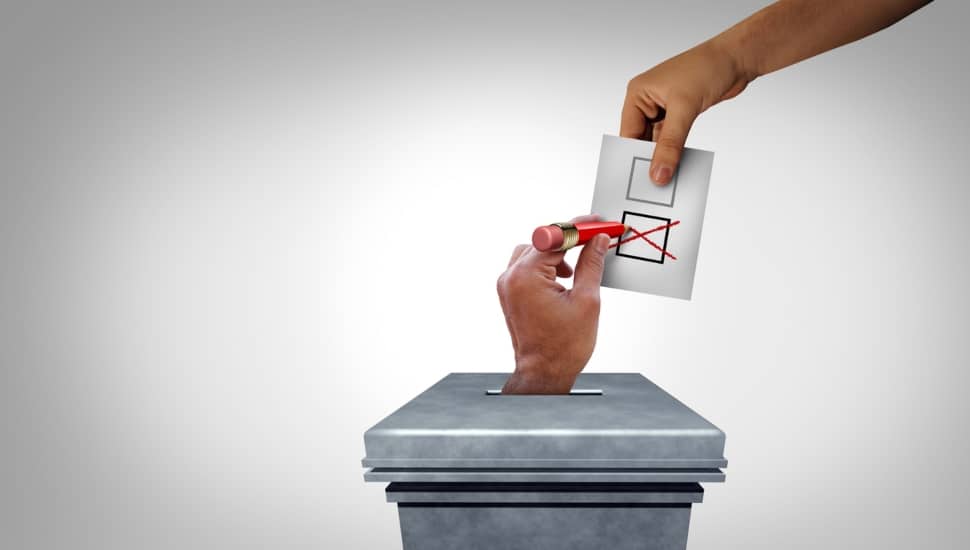 A hand reaches out of a ballot box to mark another ballot with a red X held in the hand of someone.